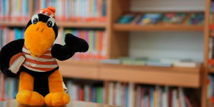 Maskottchen Rabe Fridolin in gestreiftem T-shirt vor einem Bücherregal in der Bücherei