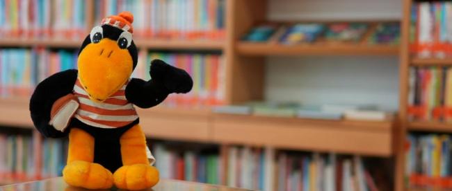 Fridolin (c) Beatrix Reese Maskottchen Rabe Fridolin in gestreiftem T-shirt vor einem Bücherregal in der Bücherei